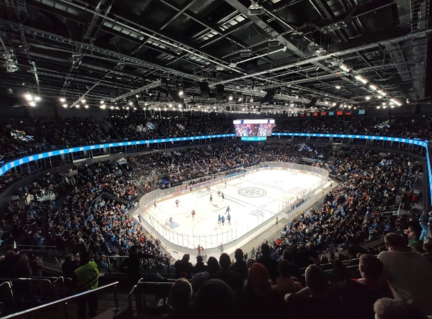 Hockey-Spiel in einer großen Arena mit Zuschauern auf den Tribünen und Personal am Boden, mit Geländern, Überwachungsbildschirmen und heller Beleuchtung, identifiziert als O2 Arena in London.