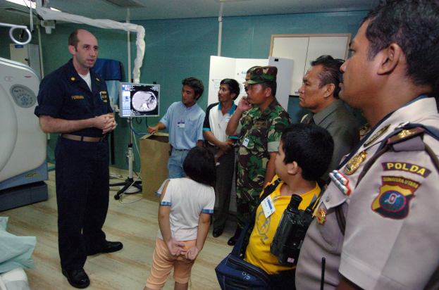 Eine Gruppe von Menschen steht um ein Krankenhauszimmer herum, ein Mann hält ein Mikrofon in der Mitte, ein Bett mit einer Decke darauf links, verschiedene medizinische Geräte, ein Röntgengerät, eine Tafel mit Text, eine Tür, eine Wand und eine Decke mit Deckenleuchten im Hintergrund.