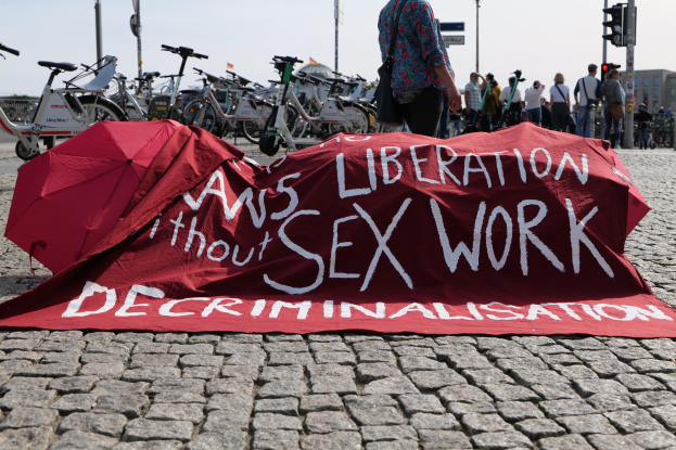 Eine Gruppe von Menschen demonstriert auf einer Stadtstraße, hält Schirme und hat ein rotes Banner mit der Aufschrift "Befreiung ohne Entkriminalisierung der Sexarbeit" auf dem Boden ausgebreitet. Fahrräder sind links geparkt, und im Hintergrund sind städtische Infrastruktur zu sehen.