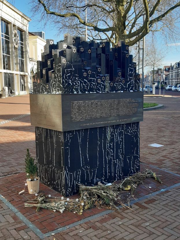Denkmal in einem Stadtplatz, das den Holocaust-Opfern gewidmet ist, mit einem Baum davor, einer Topfpflanze, verstreuten trockenen Blättern und Gebäuden, Fahrzeugen und Bäumen im Hintergrund.