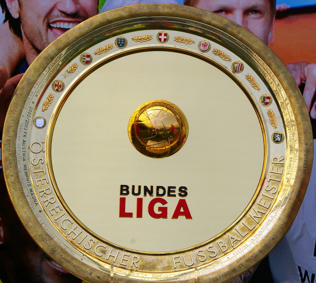 Goldener Schild mit 'Bundesliga'-Text in der Hand eines Mannes, drei Personen auf einem Banner im Hintergrund.