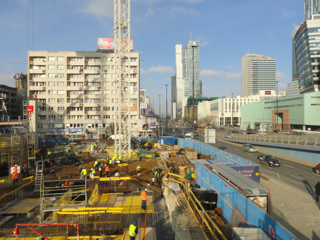 Baustelle in einer städtischen Umgebung mit Arbeitern, Fahrzeugen, Metallrahmen, Schildern, Straßenlaternen, einem Turm.