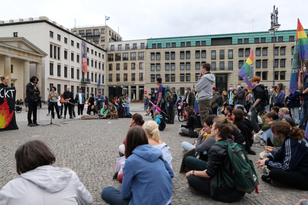 Menschen, die auf dem Boden sitzen, vor einer Menge mit Fahnen und Spruchbändern bei einer Demonstration in Berlin, mit einem Redner, einer Statue und Gebäuden im Hintergrund.