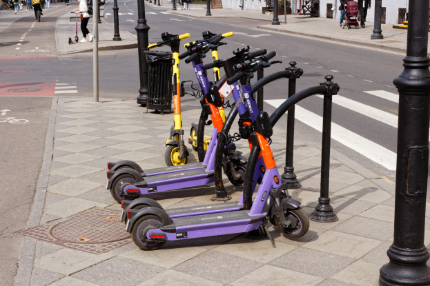 Eine Reihe von E-Scootern, die entlang einer Straße geparkt sind, mit Fußgängern, Radfahrern, einem Müllcontainer im Vordergrund und Gebäuden, Bäumen und Pflanzen im Hintergrund.