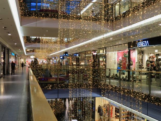 Ein festlich geschmücktes Einkaufszentrum mit Weihnachtsbeleuchtung, das Menschen zeigt, die durch die Geschäfte gehen, Schilder mit Namen der Geschäfte, Glasgeländer und von der Decke hängende Deko-Lichter.