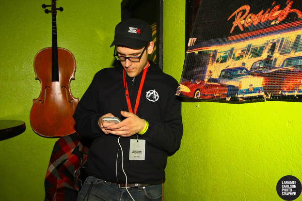 Eine Person mit Mütze und Ausweis hält ein Mobiltelefon in der Hand, daneben eine Gitarre und ein Plakat.