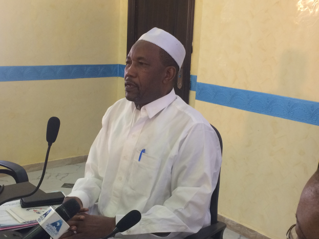 Ein Mann in weißem Anzug und Hut sitzt an einem Tisch mit Mikrofonen und spricht, während Papiere und Gegenstände auf dem Tisch zu sehen sind; rechts von ihm sitzt eine Person mit Brille, im Hintergrund eine Tür und eine Wand.