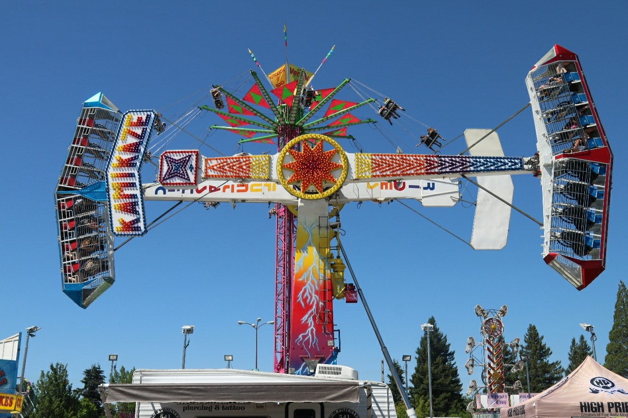 Ein Karussell mit Menschen darauf, umgeben von Zelten, Bannern, Pfählen, Lichtern und Bäumen unter einem sichtbaren Himmel.