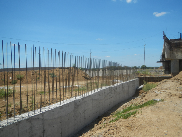 Baustelle mit einer Eisenstab-Stützmauer, einem Gebäude auf der rechten Seite und einem bewölkten Himmel im Hintergrund.