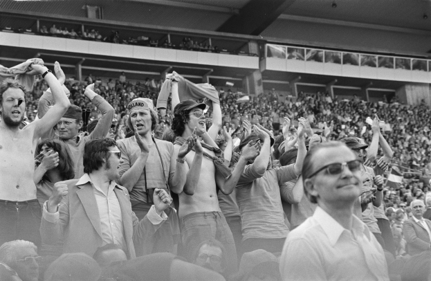 Schwarzes und weißes Foto einer vollen Tribüne mit einigen Zuschauern, die eine Brille tragen.