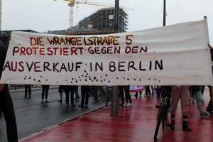 Eine Gruppe von Menschen steht auf der Straße und hält ein Schild hoch, auf dem "Die Wrangellstraße 5 protestiert gegen den Verkauf in Berlin" steht, mit Fahrrädern, Pfählen, Gebäuden und einem Kran im Hintergrund unter einem sichtbaren Himmel.