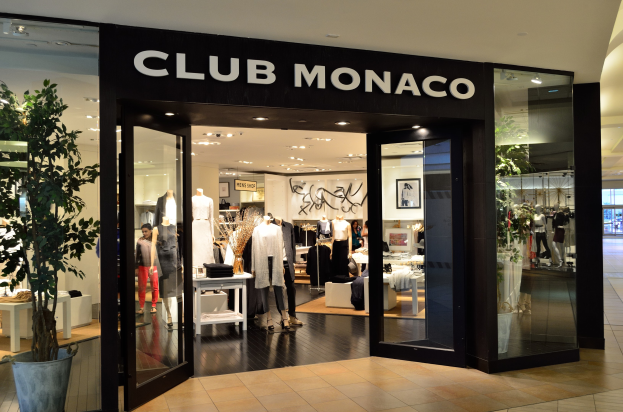 Außenansicht eines Club Monaco Geschäfts in einem Einkaufszentrum mit Glaswänden, Schaufensterpuppen in Kleidern, Auslagetischen mit Artikeln, einem Topf mit einer Pflanze und Deckenleuchten.