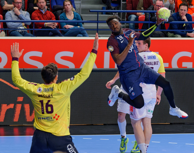 Männer beim Handballspielen auf einem Feld mit einem Ball in der Luft, Zuschauer im Hintergrund und ein Schild mit der Aufschrift "Futsal-Weltmeisterschaft 2019 - Paris Saint-Germain vs Bordeaux".