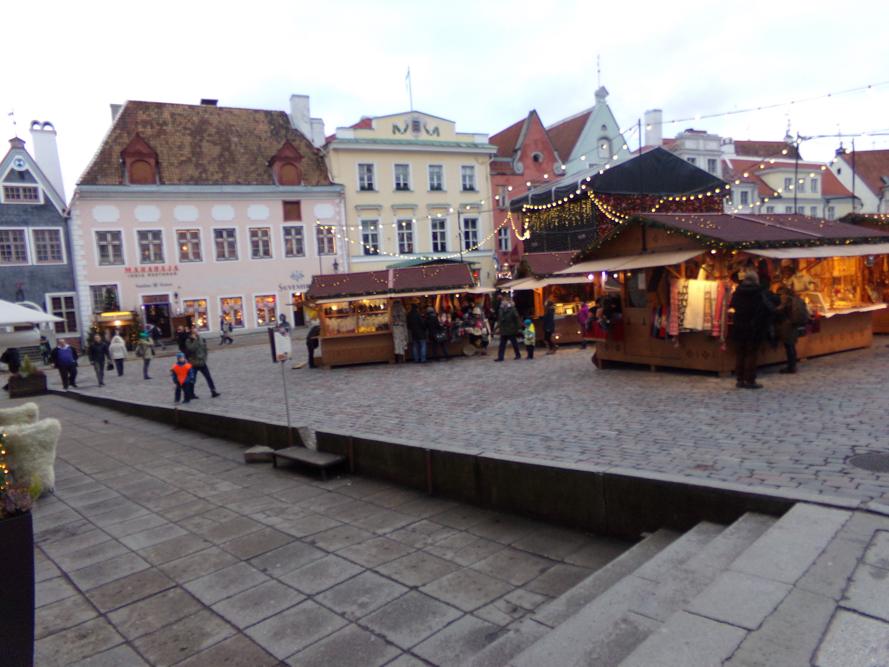 Ein geschäftiger Weihnachtsmarkt in Tallinn, Estland mit Menschen um geschmückte Stände, festliche Gebäude, bewölkten Himmel, Treppen und Topfpflanzen.