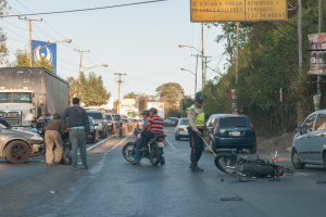 Eine Gruppe von Menschen steht um ein verunglücktes Motorrad auf der Straßenseite mit mehreren Fahrzeugen, darunter ein Lastwagen, und einem Hintergrund aus Bäumen, Masten, Lichtern und Schildern unter dem Himmel.