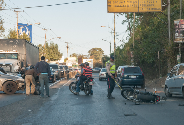 Eine Gruppe von Menschen steht um ein verunglücktes Motorrad auf der Straßenseite mit mehreren Fahrzeugen, darunter ein Lastwagen, und einem Hintergrund aus Bäumen, Masten, Lichtern und Schildern unter dem Himmel.