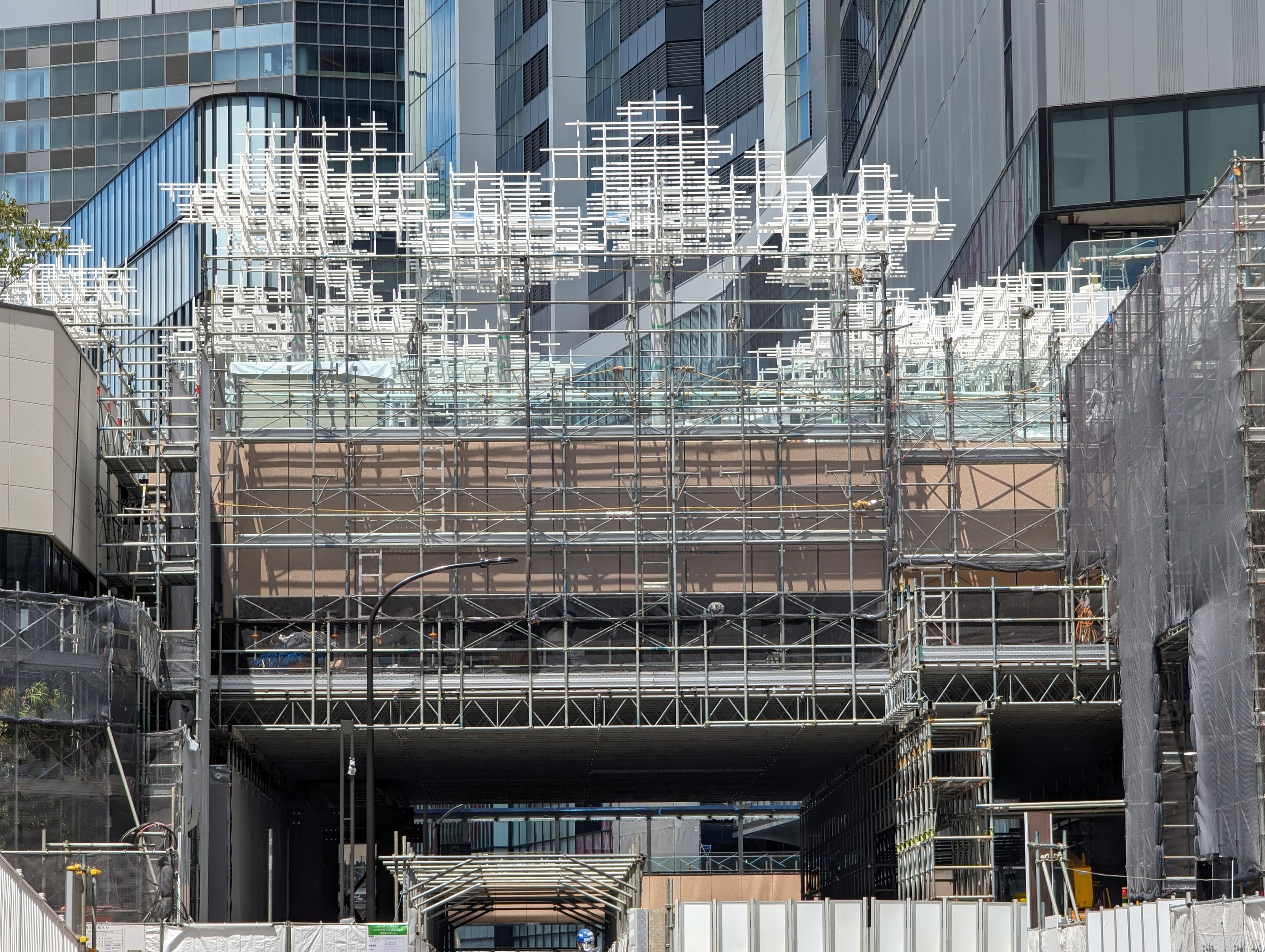 Gro├čer Bau mit Gerüst, umgeben von B├Ąumen links und einer Stra├če unten in einer urbanen Umgebung.