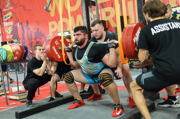 Eine Gruppe von Männern, die mit Hanteln Kniebeugen in einem Fitnessstudio machen, umgeben von Geräten und Bannern mit der Aufschrift "Powerlifting-Weltmeisterschaften 2019".