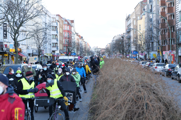 Eine große Gruppe von Menschen in Masken und Sicherheitswesten auf Fahrrädern eine von Bäumen gesäumte Straße mit Gebäuden, Laternenmasten und Texttafeln entlang, neben Fahrzeugen und trockenem Gras unter einem klaren blauen Himmel.
