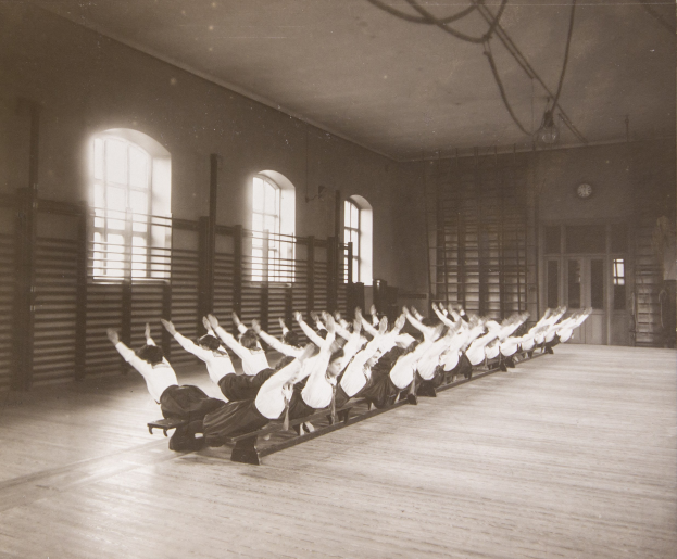 Gruppe von Menschen, die Pilates-Übungen in einem Fitnessstudio mit Fenstern und einer Uhr an der Wand im Hintergrund machen.