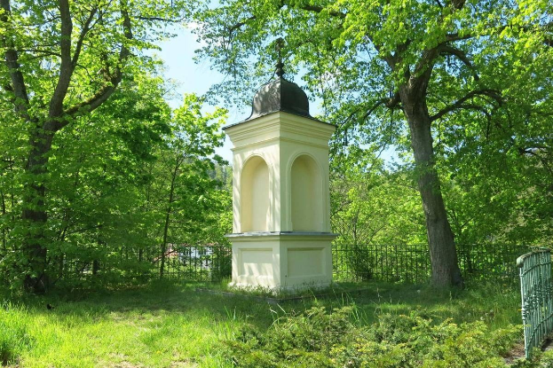 Kleines weißes Holocaust-Gedenkmal auf einem grasbewachsenen Friedhof umgeben von einem Zaun und Bäumen unter einem klaren blauen Himmel in Vilnius, Litauen.