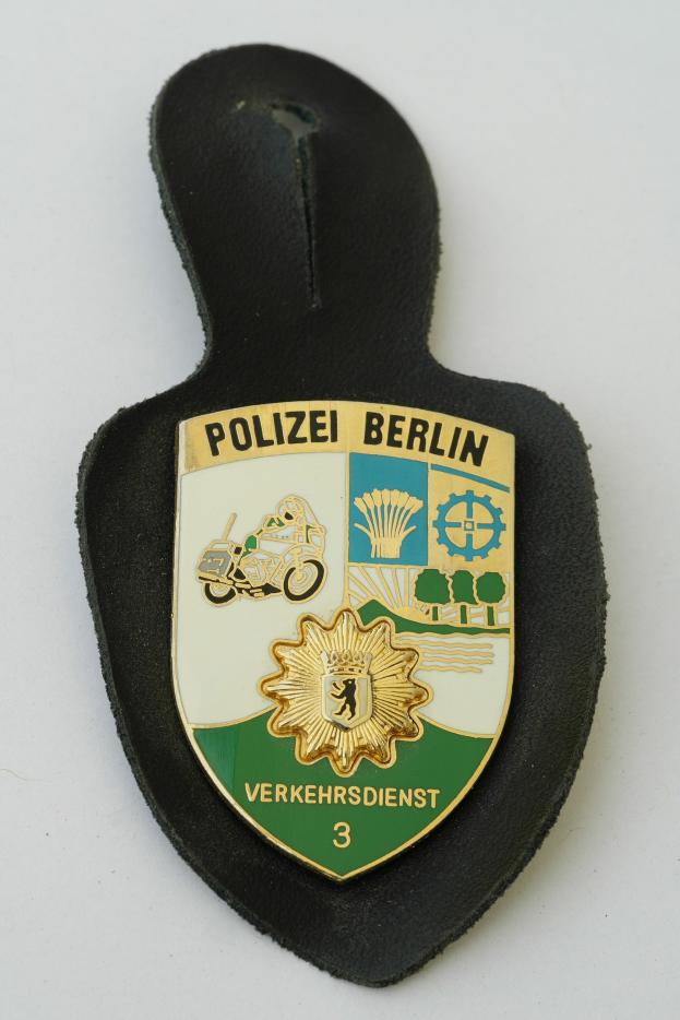 Ein schildförmiges Polizeibadge mit "Polizei Berlin" in schwarzer fetter Schrift auf einem blauen Hintergrund mit einem weißen Stern und Rand, das auf einer Oberfläche ruht.