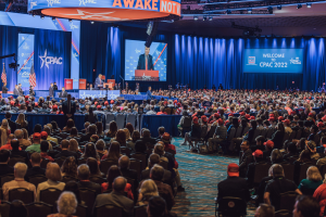 Eine große Menschenmenge sitzt auf Stühlen, die einer Bühne zugewandt sind, mit ein paar Menschen auf dem Podium und auf der linken Seite, blaue Vorhänge und ein Bildschirm im Hintergrund und eine orangefarbene Tafel oben, die das CPAC 2020-Event am 25. Oktober anzeigt.
