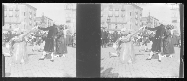 Schwarzes und weißes Foto einer Gruppe von Menschen in Kostümen, die auf der Straße tanzen, umgeben von Gebäuden, Straßenmasten und Laternen unter einem klaren Himmel.