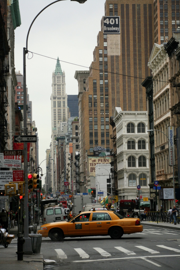 Gelbes Taxi fährt eine belebte Stadtstraße entlang mit hohen Gebäuden auf beiden Seiten, Fußgängern auf dem Gehweg, Ampeln und Texttafeln, unter einem sichtbaren Himmel.
