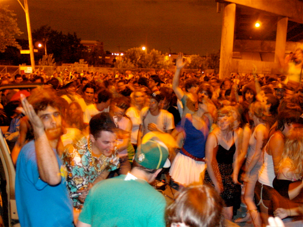 Menschen tanzen im Vordergrund eines Konzerts mit Lichtern, Masten, Bäumen und anderen Gegenständen im Hintergrund.