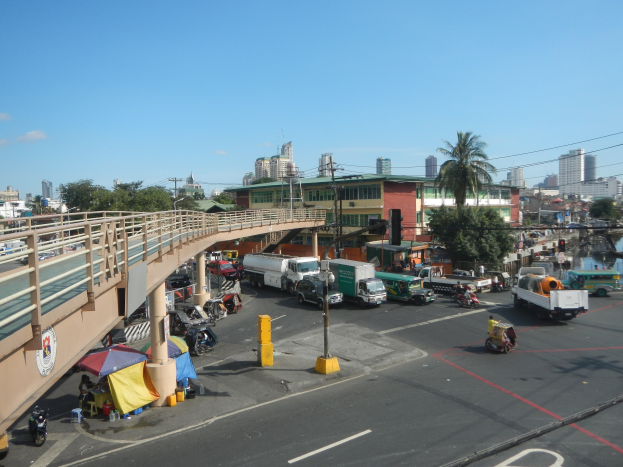 Vollgestoppte Straße mit starkem Verkehr neben einer Brücke, mit Fahrzeugen, Fußgängern, Zelten, Pfählen, Verkehrszeichen, Bäumen, Gebäuden und Wasser im Hintergrund unter einem bewölkten Himmel.