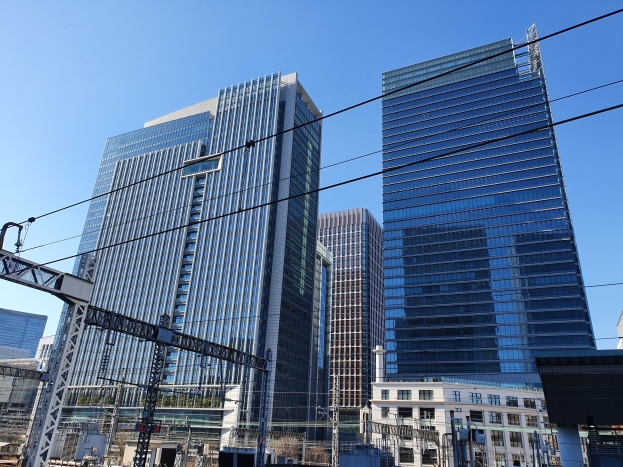Eine Stadtstraße mit hohen Gebäuden im Hintergrund, Strommasten mit Drähten, Straßenlaternen, Ampeln, Namensschilder, Bäume und einen klaren blauen Himmel.