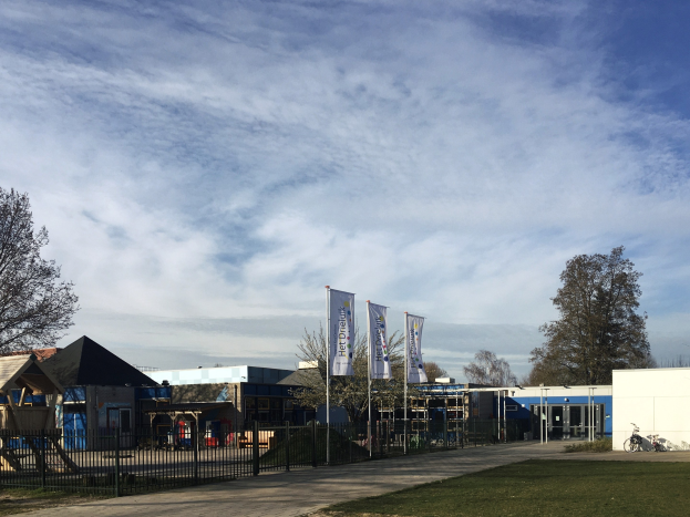 Großes Backsteingebäude mit einem grasigen Bereich davor, umgeben von einem Metallzaun, mit einem Weg, Bäumen, Bannern, einem geparkten Fahrrad, nahen Häusern und einem bewölkten Himmel, als das neue Kinder-Spielgelände an der Schule identifiziert.
