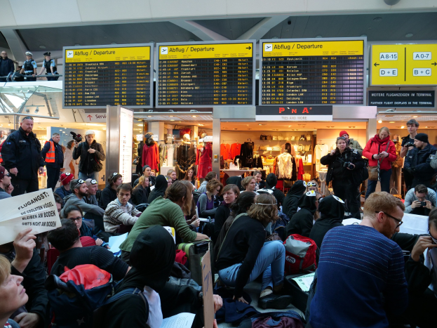 Eine große Gruppe von Menschen an einem Flughafen, einige sitzen mit Taschen und Papieren, andere stehen, mit Texttafeln, Schaufensterpuppen in Kleidern und Deckenbeleuchtung im Hintergrund, was auf eine Demonstration hindeutet.