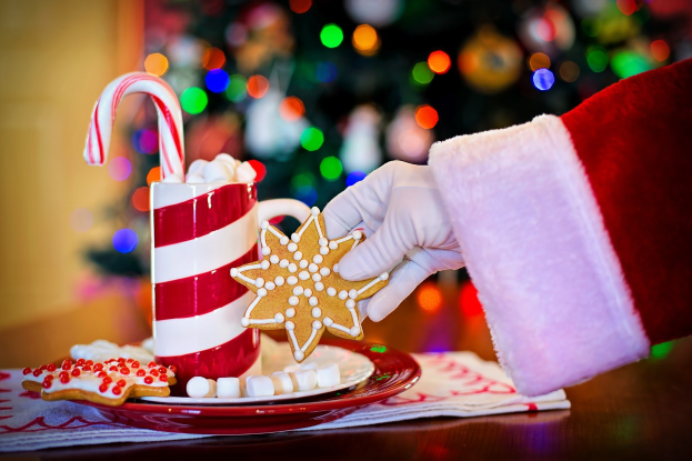 Eine Person mit einem Santa Claus Hut, die einen Becher heiße Schokolade und einen Zuckerstange hält und ein Teller mit Keksen und ein Tuch auf dem Tisch vor ihnen liegen, vor einem Weihnachtsbaum und einer Wand.