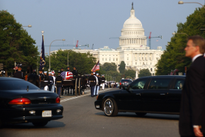 Trauerzug mit Menschen in Mützen und Fahnen vor dem Kapitolsgebäude, umgeben von Fahrzeugen, Laternenmasten, Bäumen und Gebäuden unter einem klaren Himmel.