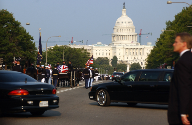 Trauerzug mit Menschen in Mützen und Fahnen vor dem Kapitolsgebäude, umgeben von Fahrzeugen, Laternenmasten, Bäumen und Gebäuden unter einem klaren Himmel.