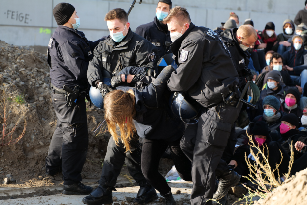 Gruppe maskierter Personen in Mützen, einige bewaffnet, umringt eine Frau, die von der Polizei eskortiert wird, mit Pflanzen, Steinen und einer protestbezogenen Wandtext im Hintergrund.