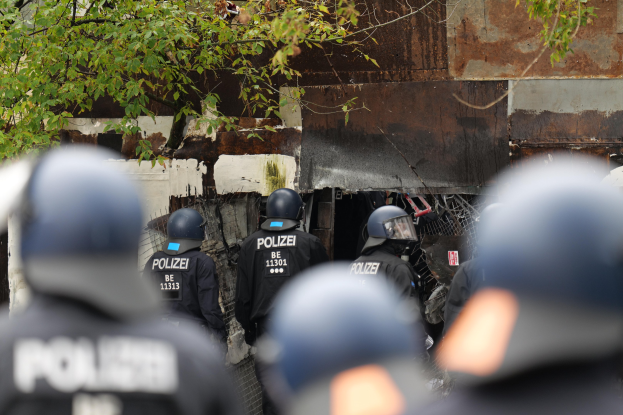 Gruppe von Polizisten in schwarzen Uniformen und Helmen, die vor einem mit Maschendrahtzaun umgebenen Geb├Ąude stehen, mit B├Ąumen im Hintergrund.