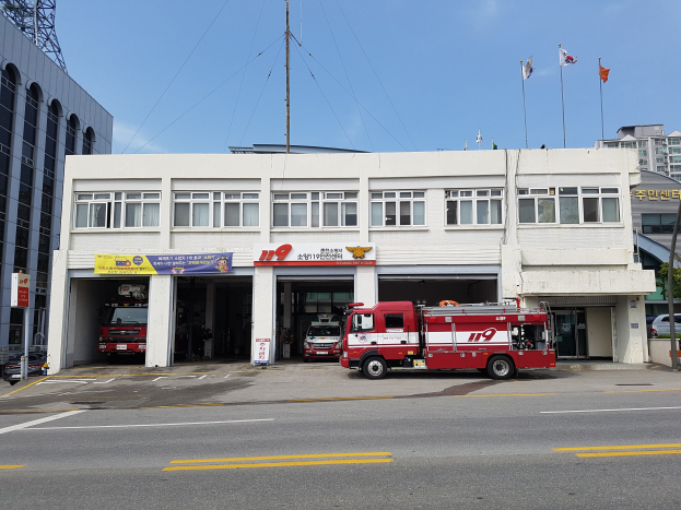 Feuerwache mit mehreren Feuerwehrautos vor der Tür, ein Gebäude mit Fenstern, Bannern und Fahnen sowie einen bewölkten Himmel mit einem Turm links.