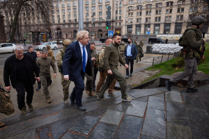 Eine Gruppe von Menschen, darunter Boris Johnson, geht eine Straße entlang neben einem Gebäude, einige tragen Helme und halten Waffen, mit Fahrzeugen, Verkehrszeichen, Bäumen und Gebäuden im Hintergrund.
