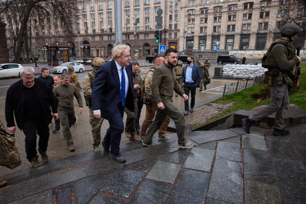 Eine Gruppe von Menschen, darunter Boris Johnson, geht eine Straße entlang neben einem Gebäude, einige tragen Helme und halten Waffen, mit Fahrzeugen, Verkehrszeichen, Bäumen und Gebäuden im Hintergrund.