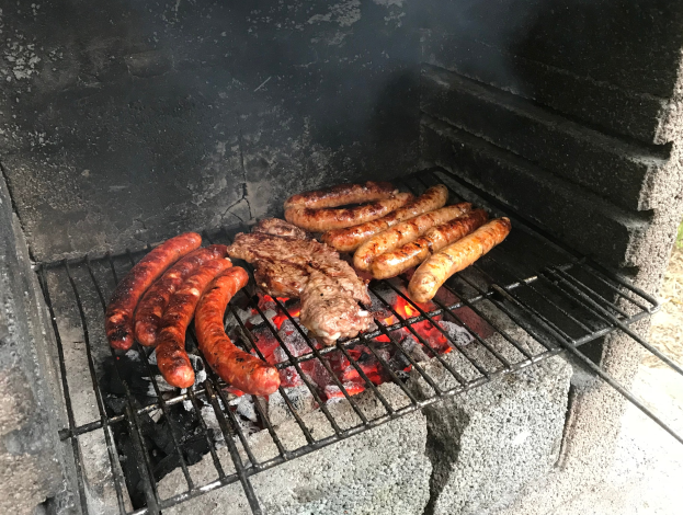 Ein Grill mit Würstchen und Fleisch darauf, umgeben von Kohle und Feuer, umgeben von Gras.