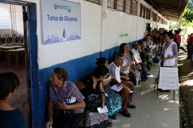 Eine Gruppe von Menschen sitzt auf Bänken vor einem Gebäude mit einer Texttafel an der Wand und einer Tür links, die auf eine Wahlurne in Guadalajara, Mexiko, warten.
