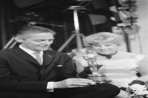 Ein Mann in einem schwarzen Blazer, einem weißen Hemd und einer Krawatte hölt eine Oscar-Statue, waehrend er neben einer Frau in formeller Kleidung sitzt, die einen Blumenstrauß hölt, mit Hintergrundlichtern, die die schwarz-weiße Szenerie erhellen.