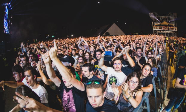 Eine große Menschenmenge auf einem Musikfestival mit Menschen, die ihre Hände heben, einige sitzen und andere stehen, in der Nähe eines Metallzauns mit Gebäuden und Lichtern im Hintergrund.