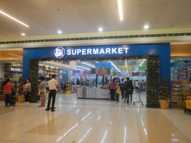 Eine Gruppe von Menschen steht vor einem Supermarkt, einige halten Taschen, mit einem Einkaufswagen, Topfpflanze, Gestellen mit Gegenständen, Schildern und einer beleuchteten Decke im Hintergrund.