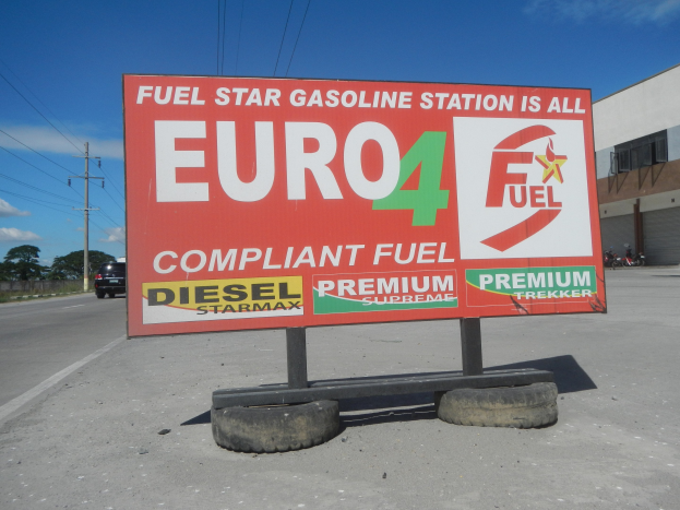 Eine Tankstelle mit einem "Euro4 Fuel"-Schild im Vordergrund, Fahrzeuge auf der Straße, Strommasten mit Drähten, Bäume, ein Gebäude auf der rechten Seite und sichtbarer Himmel.