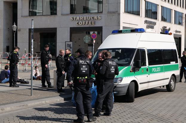 Eine Gruppe von Polizisten steht vor einem Starbucks-Café, rechts steht ein Van und links sitzen und stehen ein paar Menschen; im Hintergrund sind ein Gebäude, ein Schild, ein Laternenpfahl und ein Zaun zu sehen.