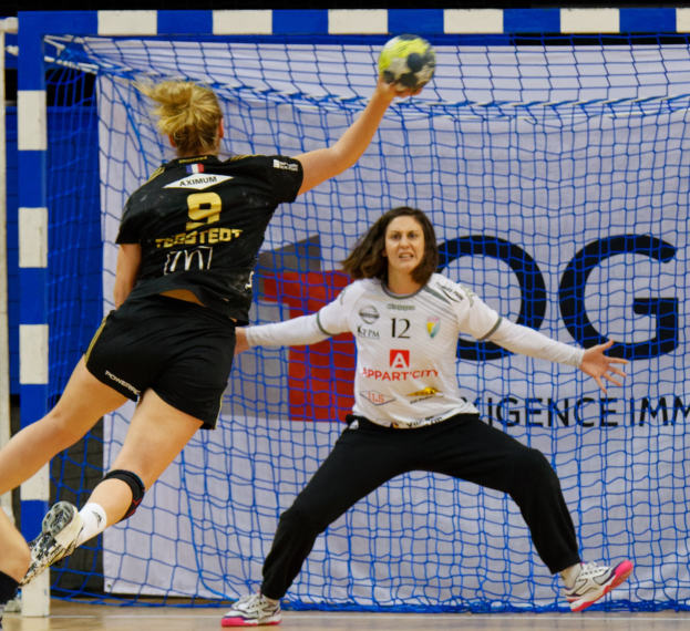 Zwei Frauen beim Handballspiel vor einem Tor, eine hält den Ball, im Hintergrund ein Netz und eine Tafel mit Text.